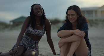 Movie still from “Along for the Ride” (2022), directed by Sofia Alvarez – Two women sitting on the beach smiling for the camera; Medium shot, Over the shoulder angle
