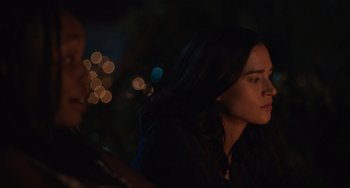 Movie still from “Along for the Ride” (2022), directed by Sofia Alvarez – A woman sitting in front of a tree with lights in the background; Close Up shot, Over the shoulder angle