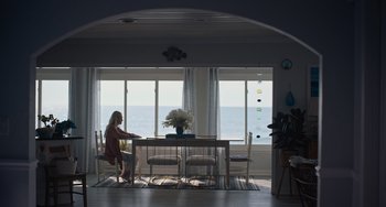 Movie still from “Along for the Ride” (2022), directed by Sofia Alvarez – A woman sitting at a dining room table in front of the ocean; Wide shot, Low angle