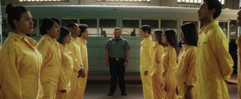 Movie still from “American Carnage” (2022), directed by Diego Hallivis – A group of people in yellow suits standing in front of a bus; Wide shot, Low angle