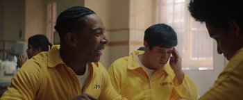 Movie still from “American Carnage” (2022), directed by Diego Hallivis – Two young men in yellow prison uniforms are smiling; Medium shot, Over the shoulder angle
