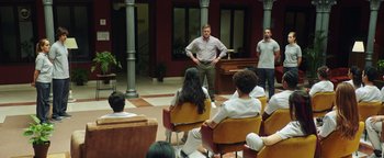 Movie still from “American Carnage” (2022), directed by Diego Hallivis – A group of people sitting in a room with a man standing in front of them; Wide shot, Over the shoulder angle