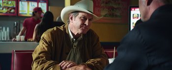 Movie still from “American Carnage” (2022), directed by Diego Hallivis – A man wearing a white cowboy hat sitting at a table; Medium shot, Over the shoulder angle