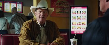 Movie still from “American Carnage” (2022), directed by Diego Hallivis – An older man wearing a cowboy hat sitting at a table in front of an advertisement; Medium shot, Low angle