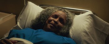 Movie still from “American Carnage” (2022), directed by Diego Hallivis – An older woman laying in a hospital bed smiling; Close Up shot, Overhead angle