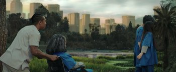 Movie still from “American Carnage” (2022), directed by Diego Hallivis – An older woman sitting on a park bench looking out at a city skyline; Wide shot, Over the shoulder angle