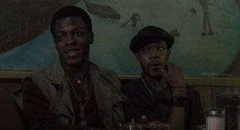 Movie still from “American Gangster” (2007), directed by Ridley Scott – Two men sitting at a table in front of a painting; Close Up shot, Over the shoulder angle