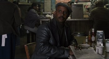 Movie still from “American Gangster” (2007), directed by Ridley Scott – A man sitting at a table in front of a newspaper; Medium shot, Over the shoulder angle