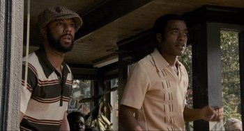 Movie still from “American Gangster” (2007), directed by Ridley Scott – Two men standing next to each other in front of a building; Medium shot, Low angle