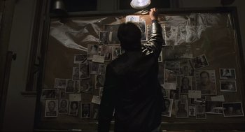 Movie still from “American Gangster” (2007), directed by Ridley Scott – A person standing in front of a wall covered in pictures; Medium shot, High angle