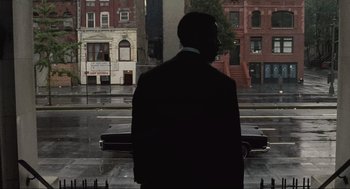 Movie still from “American Gangster” (2007), directed by Ridley Scott – A man standing in front of a building in the rain; Medium shot, Over the shoulder angle