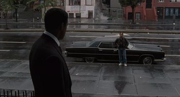 Movie still from “American Gangster” (2007), directed by Ridley Scott – A man standing on the side of the road next to a car; Wide shot, Over the shoulder angle