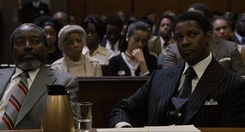 Movie still from “American Gangster” (2007), directed by Ridley Scott – A man sitting in front of a group of people in a courtroom; Medium shot, Over the shoulder angle