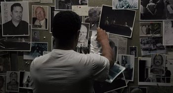 Movie still from “American Gangster” (2007), directed by Ridley Scott – A man standing in front of a wall covered in photos; Medium shot, Over the shoulder angle