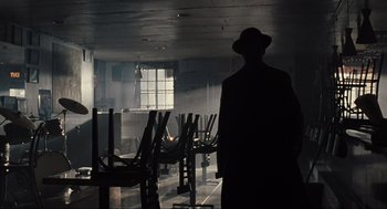 Movie still from “American Gangster” (2007), directed by Ridley Scott – A man standing in front of a group of wooden chairs; Wide shot, Over the shoulder angle
