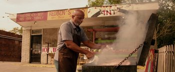 Movie still from “The Hate U Give” (2018), directed by George Tillman Jr. – A man cooking food on an outdoor grill; Wide shot, Low angle