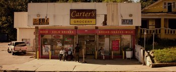 Movie still from “The Hate U Give” (2018), directed by George Tillman Jr. – Three people sitting outside of a grocery store on the sidewalk; Extreme Wide shot, Low angle