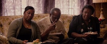 Movie still from “The Hate U Give” (2018), directed by George Tillman Jr. – An older man sitting on a couch eating a sandwich; Medium shot, High angle