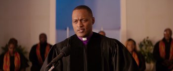 Movie still from “The Hate U Give” (2018), directed by George Tillman Jr. – A man wearing a black robe and a purple clerical collar; Close Up shot, Low angle
