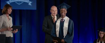 Movie still from “The Hate U Give” (2018), directed by George Tillman Jr. – A man standing next to another man in a cap and gown; Medium shot, Over the shoulder angle