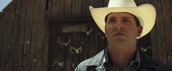 Movie still from “American Sniper” (2014), directed by Clint Eastwood – A man wearing a cowboy hat standing in front of antlers; Close Up shot, Low angle