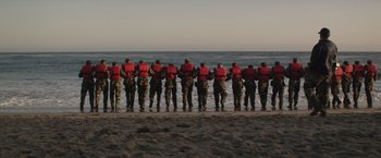Movie still from “American Sniper” (2014), directed by Clint Eastwood – A group of men standing on top of a sandy beach next to the ocean; Wide shot, Low angle