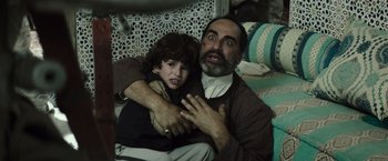 Movie still from “American Sniper” (2014), directed by Clint Eastwood – An older man holding a young boy in his arms; Medium shot, Over the shoulder angle