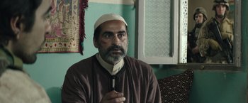 Movie still from “American Sniper” (2014), directed by Clint Eastwood – A man with a beard and a white hat is holding his hands together; Close Up shot, Over the shoulder angle