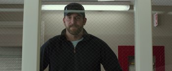 Movie still from “American Sniper” (2014), directed by Clint Eastwood – A man wearing a hat standing in front of a fence; Medium shot, Over the shoulder angle