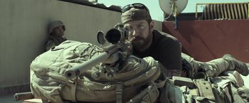 Movie still from “American Sniper” (2014), directed by Clint Eastwood – A man with a beard and a hat is holding a rifle; Medium shot, Over the shoulder angle