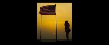 Movie still from “American Sniper” (2014), directed by Clint Eastwood – A man standing next to a flag on a pole; Wide shot, Low angle