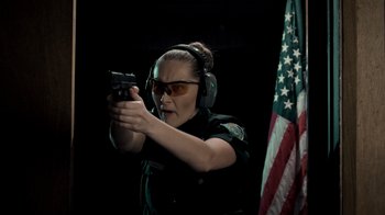 Movie still from “Amityville Uprising” (2022), directed by Thomas J. Churchill – A female police officer holding a gun in front of an american flag; Medium shot, Low angle