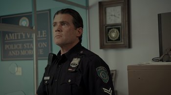 Movie still from “Amityville Uprising” (2022), directed by Thomas J. Churchill – A police officer is standing in front of a clock; Medium shot, Low angle