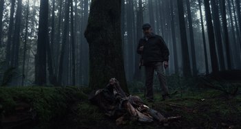 Movie still from “Antlers” (2021), directed by Scott Cooper – A man standing next to a tree in the woods; Wide shot, Low angle