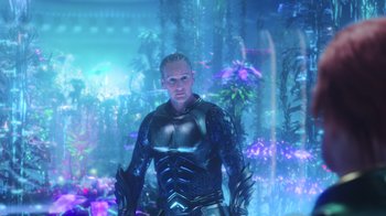 Movie still from “Aquaman” (2018), directed by James Wan – A man in a blue and black outfit standing in front of some plants; Medium shot, Over the shoulder angle