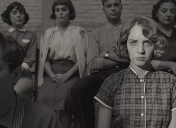 Movie still from “Asteroid City” (2023), directed by Wes Anderson – A group of people sitting in a room with a woman in front of a brick wall; Medium shot, Over the shoulder angle