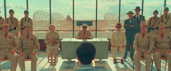 Movie still from “Asteroid City” (2023), directed by Wes Anderson – A group of people sitting at a table in front of a window; Wide shot, Over the shoulder angle
