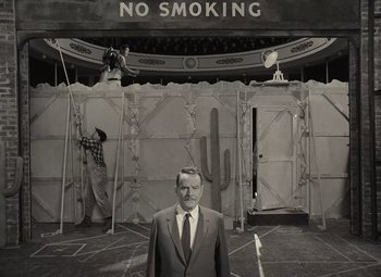Movie still from “Asteroid City” (2023), directed by Wes Anderson – A man in a suit and tie standing in front of a no smoking sign; Wide shot, Low angle
