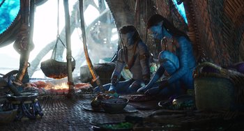 Movie still from “Avatar: The Way of Water” (2022), directed by James Cameron – A couple of people sitting on the ground near a fire pit; Wide shot, Over the shoulder angle
