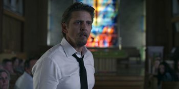 Movie still from “Awake” (2021), directed by Mark Raso – A man wearing a white shirt and a black tie in front of a stained glass window; Medium shot, Over the shoulder angle