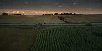 Movie still from “Awake” (2021), directed by Mark Raso – A car driving down a road near a field of crops; Extreme Wide shot, High angle