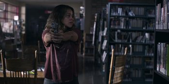 Movie still from “Awake” (2021), directed by Mark Raso – A woman holding a gun in a library; Medium shot, Over the shoulder angle