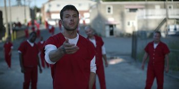 Movie still from “Awake” (2021), directed by Mark Raso – A man in a red prison uniform points to the ground; Medium shot, Over the shoulder angle