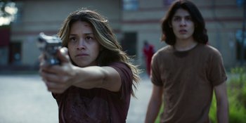 Movie still from “Awake” (2021), directed by Mark Raso – A young woman points a gun at the camera while a young man walks behind her; Medium shot, Over the shoulder angle