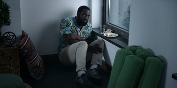 Movie still from “Awake” (2021), directed by Mark Raso – A man sitting on a window sill in front of a plate of food; Medium shot, High angle