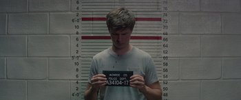 Movie still from “Baby Driver” (2017), directed by Edgar Wright – A man holding up a police department sign in front of a wall; Medium shot, Low angle