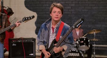 Movie still from “Back to the Future” (1985), directed by Robert Zemeckis – A young man holding a guitar in front of a group of people; Medium shot, Low angle