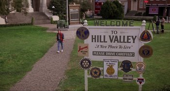 Movie still from “Back to the Future” (1985), directed by Robert Zemeckis – A woman walking down a path in front of a welcome to hill valley sign; Extreme Wide shot, High angle