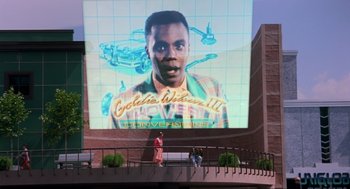 Movie still from “Back to the Future Part II” (1989), directed by Robert Zemeckis – People are standing on a balcony in front of a large screen with an image of a black man; Extreme Wide shot, High angle
