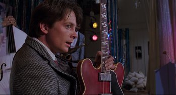 Movie still from “Back to the Future Part II” (1989), directed by Robert Zemeckis – A young man holding a guitar in a room with lights; Medium shot, Over the shoulder angle